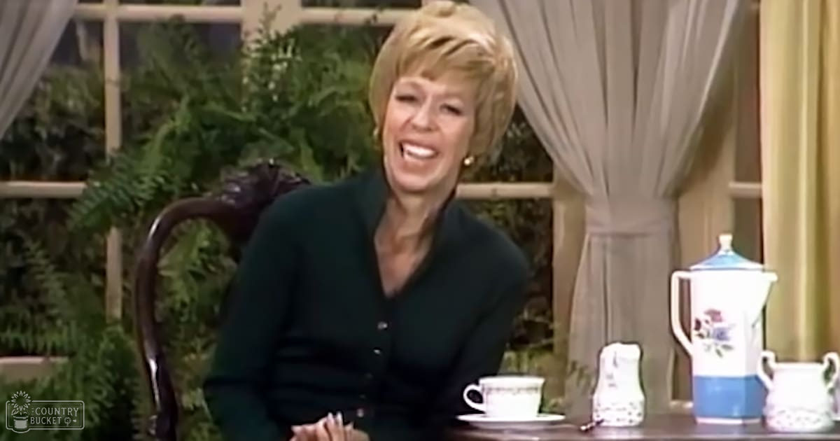 A middle-aged woman in a relaxed, comfortable fit dark green cardigan with buttons down the front. The setting appears to be indoors, possibly a home or studio, with natural light coming from outside and plants suggesting it might be spring or summer.