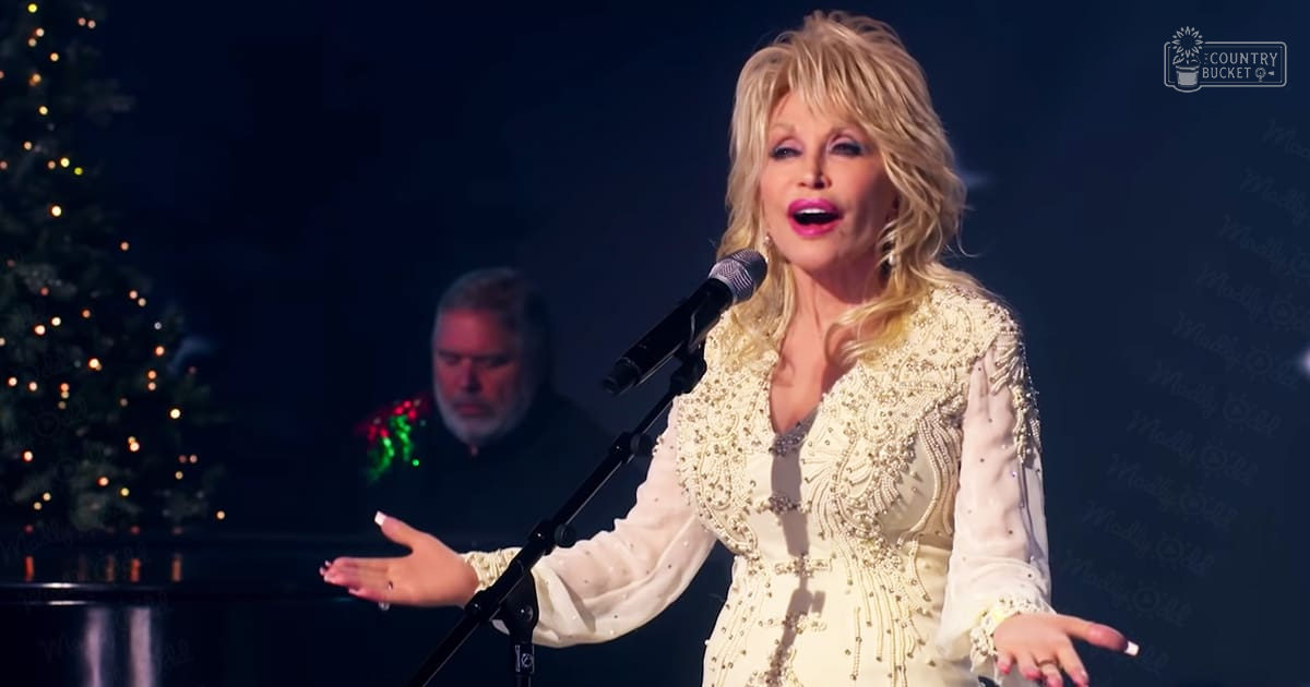 A female performer sings on stage in a Christmas-themed setting, wearing an intricate white outfit with beadwork and fringe details. A middle-aged male is visible in the background, dressed casually.