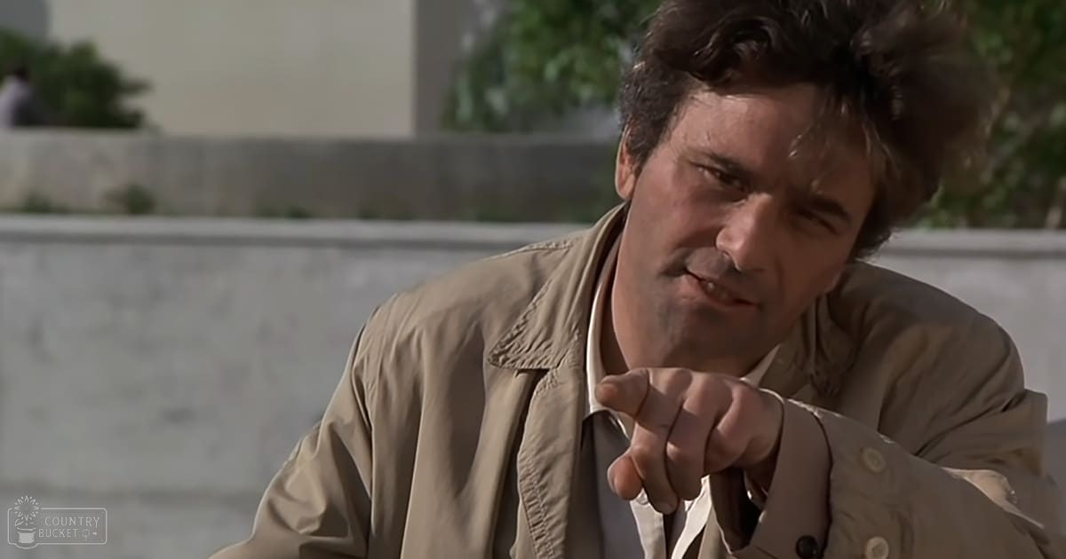 A middle-aged man wearing a beige trench coat with a light-colored shirt underneath, standing outdoors near a stone wall and some greenery.