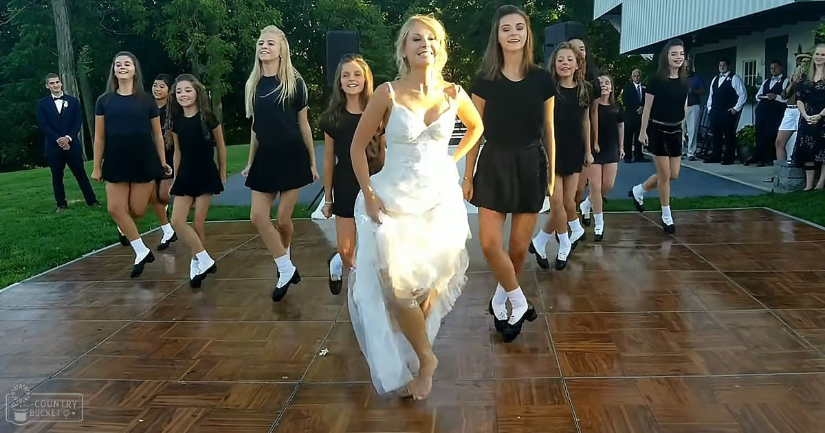 A bride dancing with a group of young women at an outdoor wedding reception, surrounded by greenery and white siding buildings in the background. The scene is bathed in late afternoon sunlight.