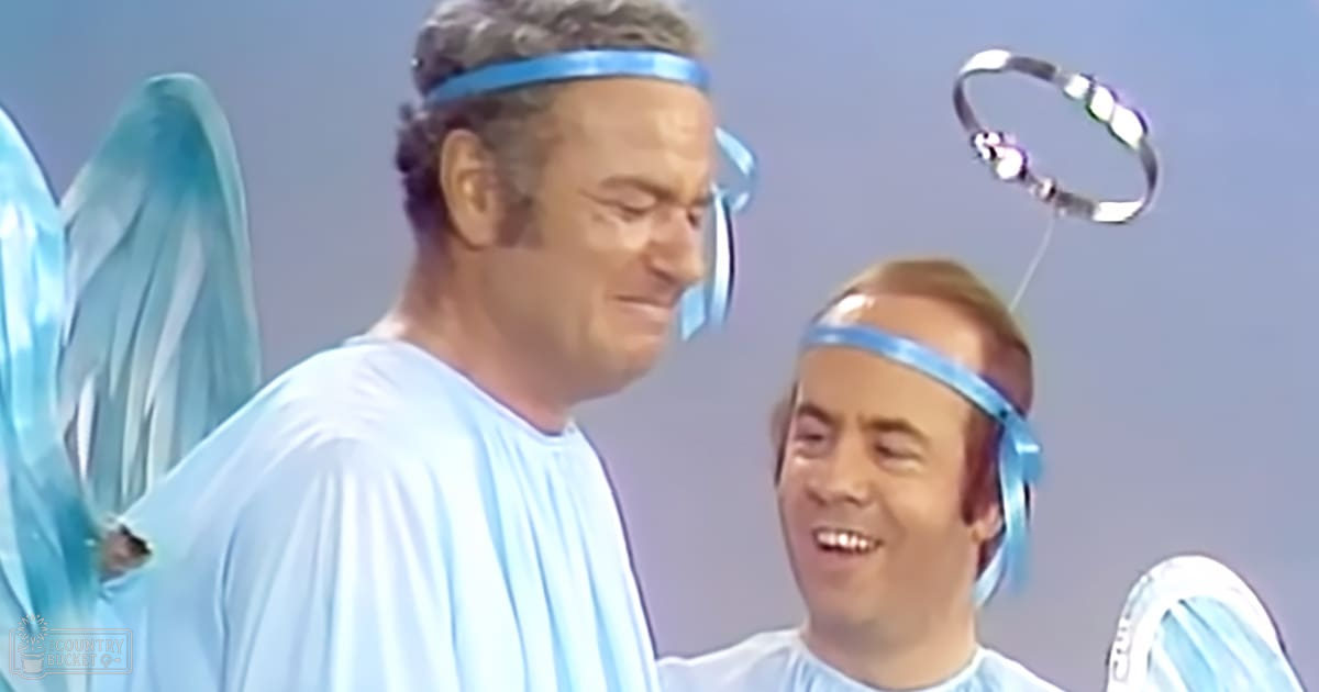 A theatrical performance scene with two older adult males dressed as angelic figures in light blue robes and translucent wings, set against a muted grayish-blue backdrop indoors.