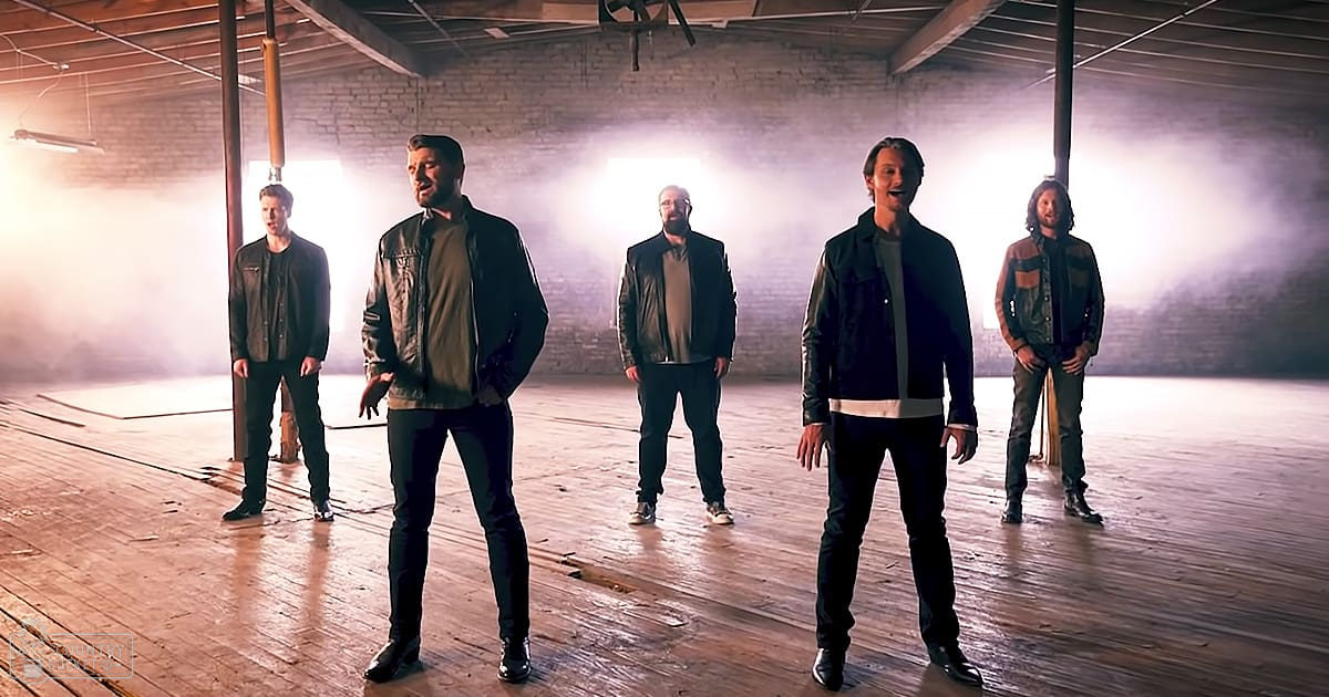 Five adult males stand together in a bright, diffused-lit industrial space with wooden floors and exposed brick walls; earthy tones dominate the scene.