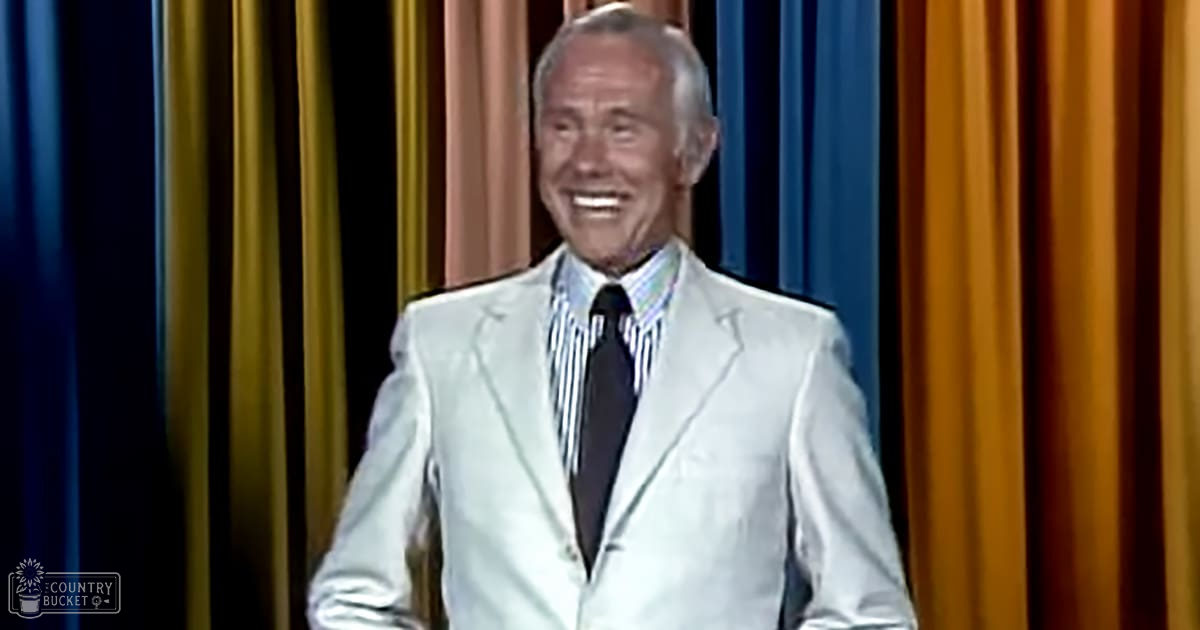 An older adult male wearing a white tailored suit with a striped shirt, standing on stage in front of curtains under artificial evening lighting.