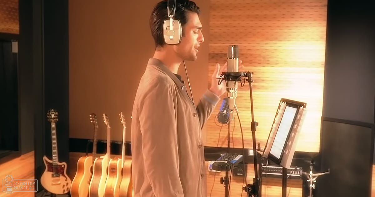 A young adult male singer in a recording studio, wearing headphones and a light brown button-up shirt, surrounded by musical equipment under natural daylight.
