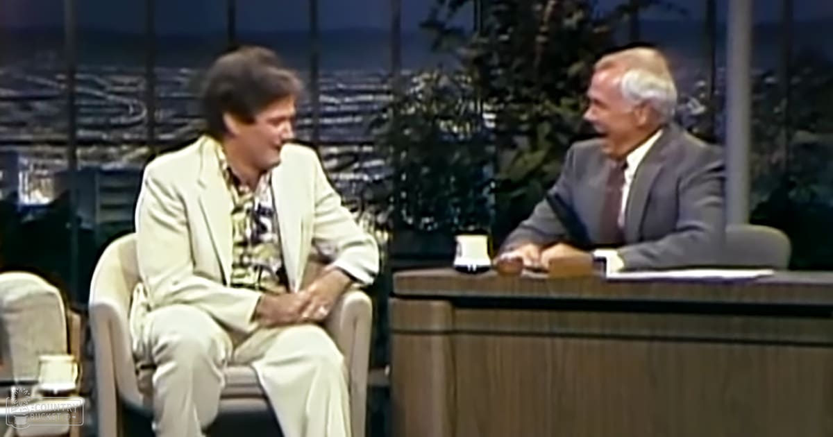 Adult male interviewee in a light-colored suit jacket and patterned shirt sits across from another adult male in a dark suit with a white shirt and patterned tie, in an indoor studio setting with large windows showing cityscape at dusk.