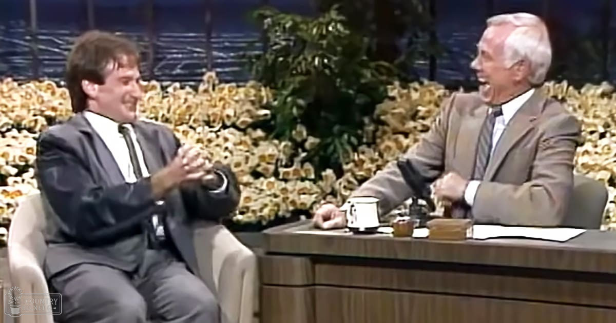 Two adult males seated in a talk show setting, one wearing a dark suit and white shirt with black tie, the other older male in a beige suit jacket over a white collared shirt with striped tie, both on a light-colored chair and wooden desk, respectively.