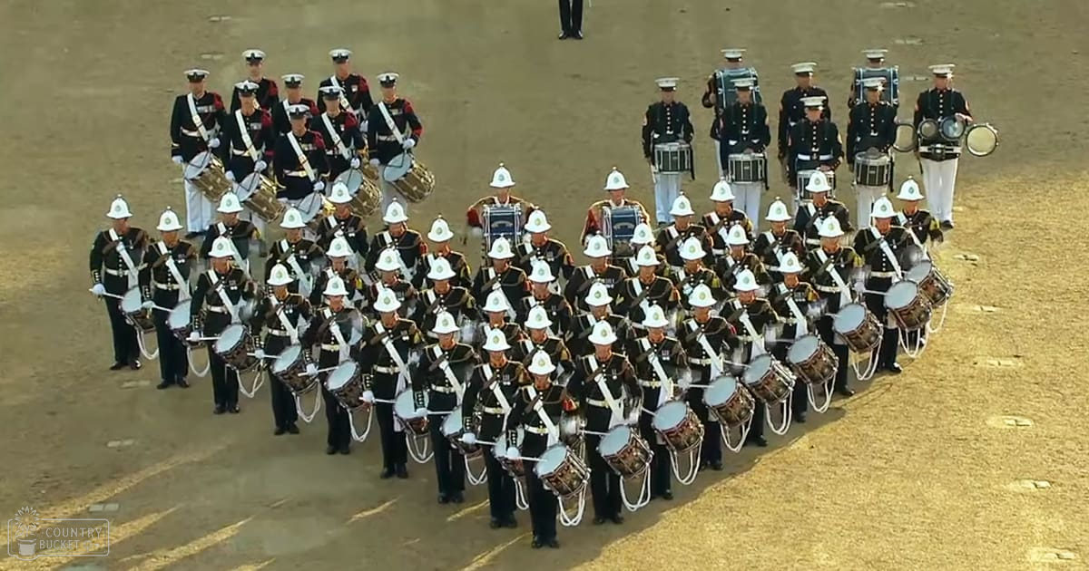 Adult male soldiers in formal military uniforms with white belts and hats marching on an open field under bright daylight, sandy terrain visible beneath them