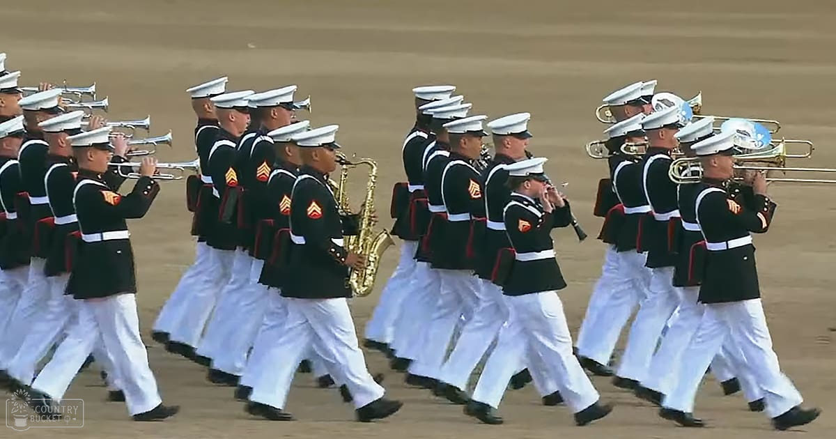 A marching band in formal military attire performs at an outdoor military parade on sandy ground under natural daylight, likely spring or summer.