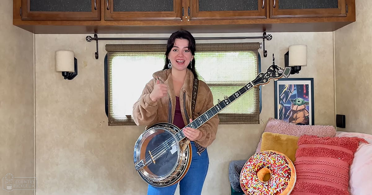 A young adult female, likely in her 20s, wears a brown fur-lined jacket over maroon top and blue jeans. She holds a banjo with black neck and silver body, near a large donut with colorful sprinkles on an RV interior. Wall sconces are mounted on either side of the window.