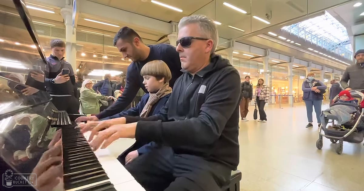 Adult male wearing sunglasses, dark jacket, and light-colored pants playing piano with two children in an indoor public space, likely a train station or airport.