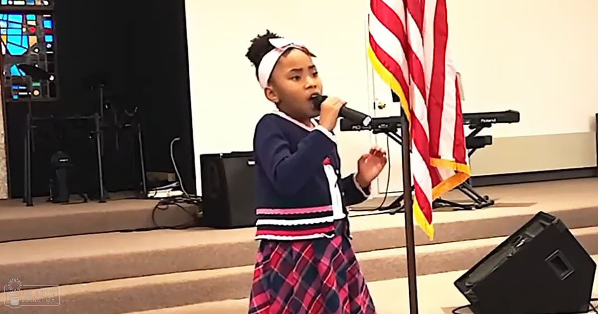 A young female child wearing a dark blue cardigan with white trim, a plaid skirt in red, black, and navy, and a white headband stands on stage during an event or performance within a church-like setting. Musical equipment and an American flag are visible behind her.