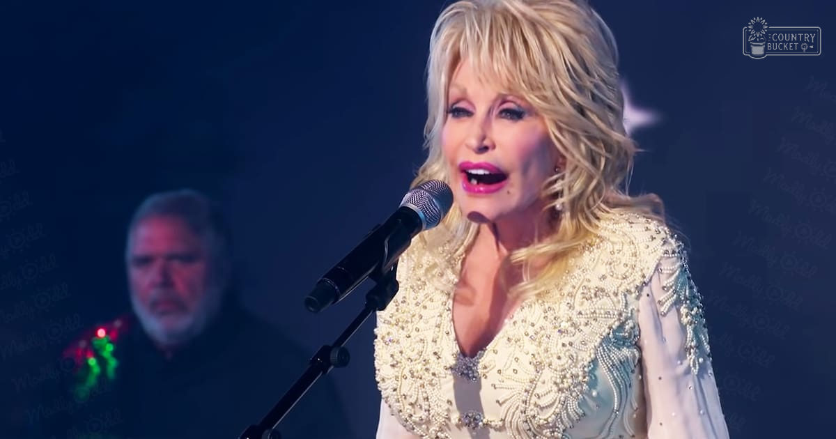 An adult female singer in a cream-colored, intricately beaded and embroidered outfit performs at an indoor concert or performance setting. A middle-aged man in a dark shirt with red-green patterned design stands in the background.