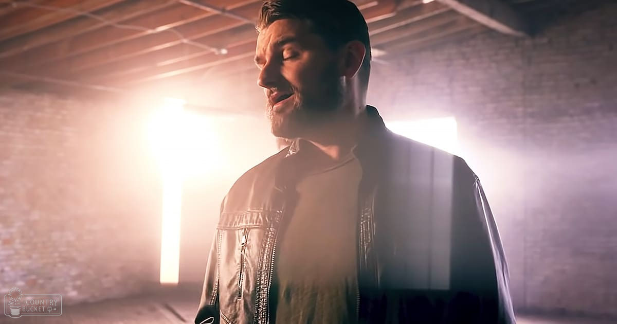 Young adult male wearing a relaxed fit, collarless white t-shirt with long sleeves in an industrial setting with exposed brick walls and metal beams; bright light sources create dramatic effect.