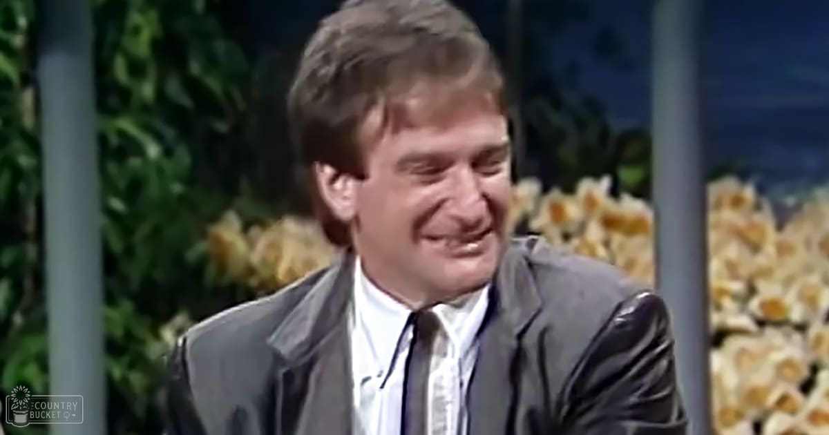 An adult male in a studio setting, wearing a dark gray suit jacket over a white dress shirt and black tie with subtle stripes, against a backdrop of greenery and yellow flowers.