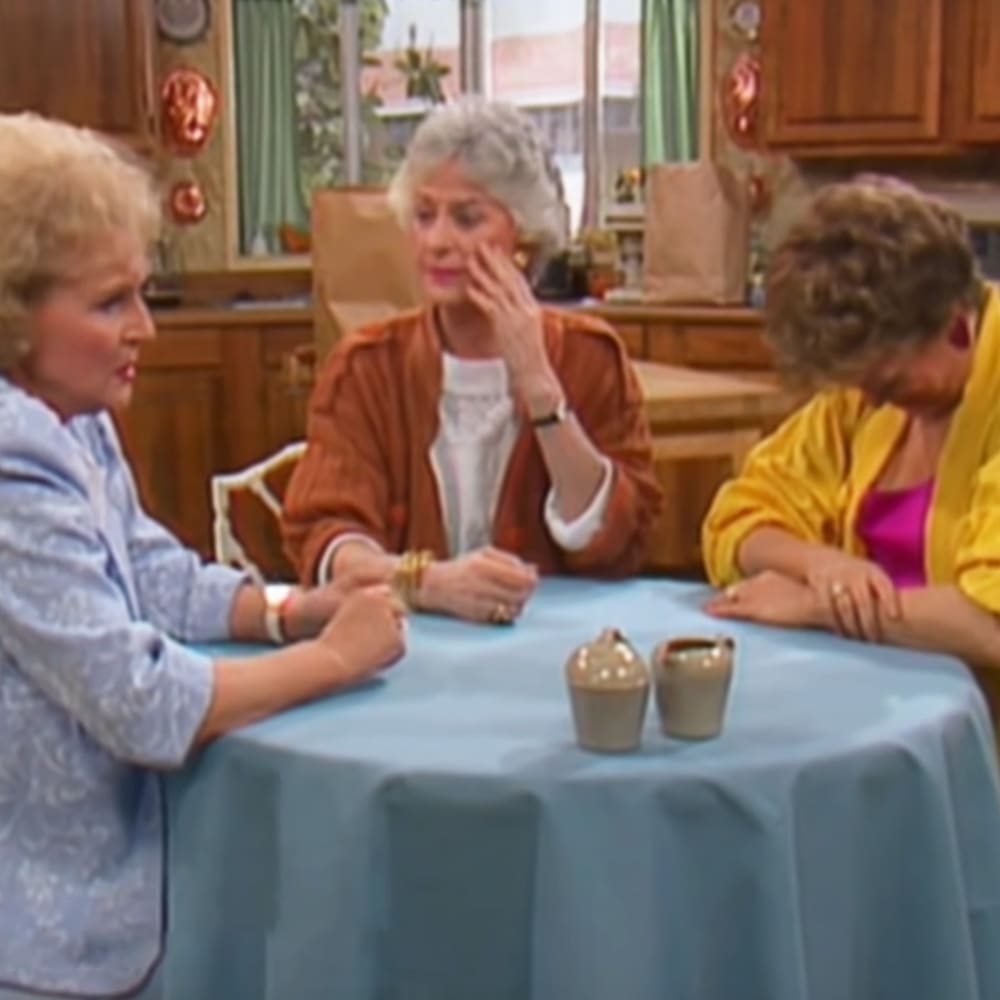 A middle-aged woman wearing a light blue patterned blouse interacts with two other women in an indoor kitchen setting; one wears an orange cardigan over a white top, and the third is dressed in a yellow jacket over pink. Focus on small ceramic containers on a table covered with a light blue tablecloth.