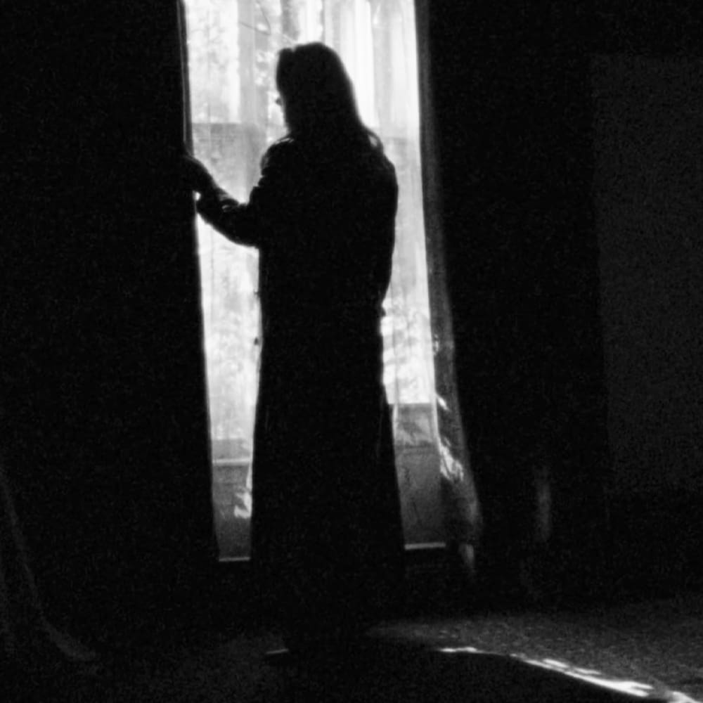 Young adult female wearing a loose, flowing long dress or robe in an indoor setting with natural light coming through large windows suggesting daytime and possibly autumn.