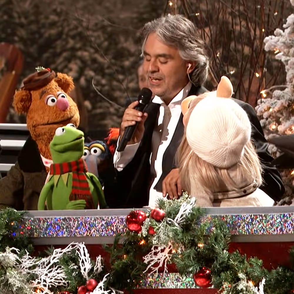 Andrea Bocelli performing with Muppets in a festive Christmas setting, wearing a black tuxedo; Fozzie Bear as a child male in a brown fur costume, Kermit the Frog as a child male in a green fur and red scarf, Miss Piggy as an adult female in a white fur coat and beige hat.