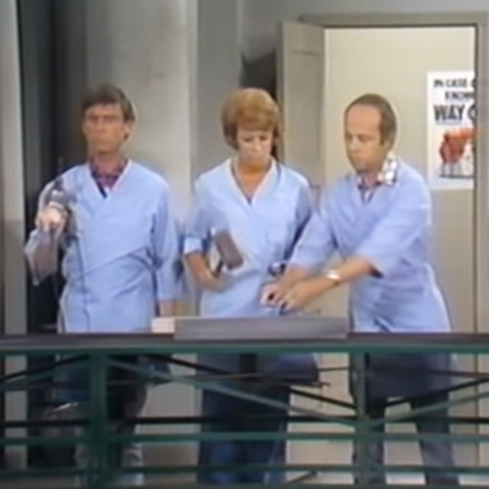 Three adult individuals wearing light blue scrub tops, working together in what appears to be a medical or hospital setting.
