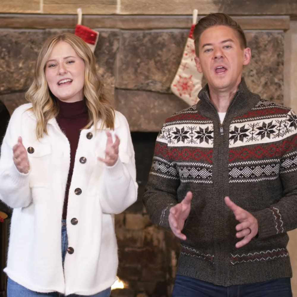 A couple in front of a stone fireplace with festive stockings hanging, suggesting a cozy holiday setting.