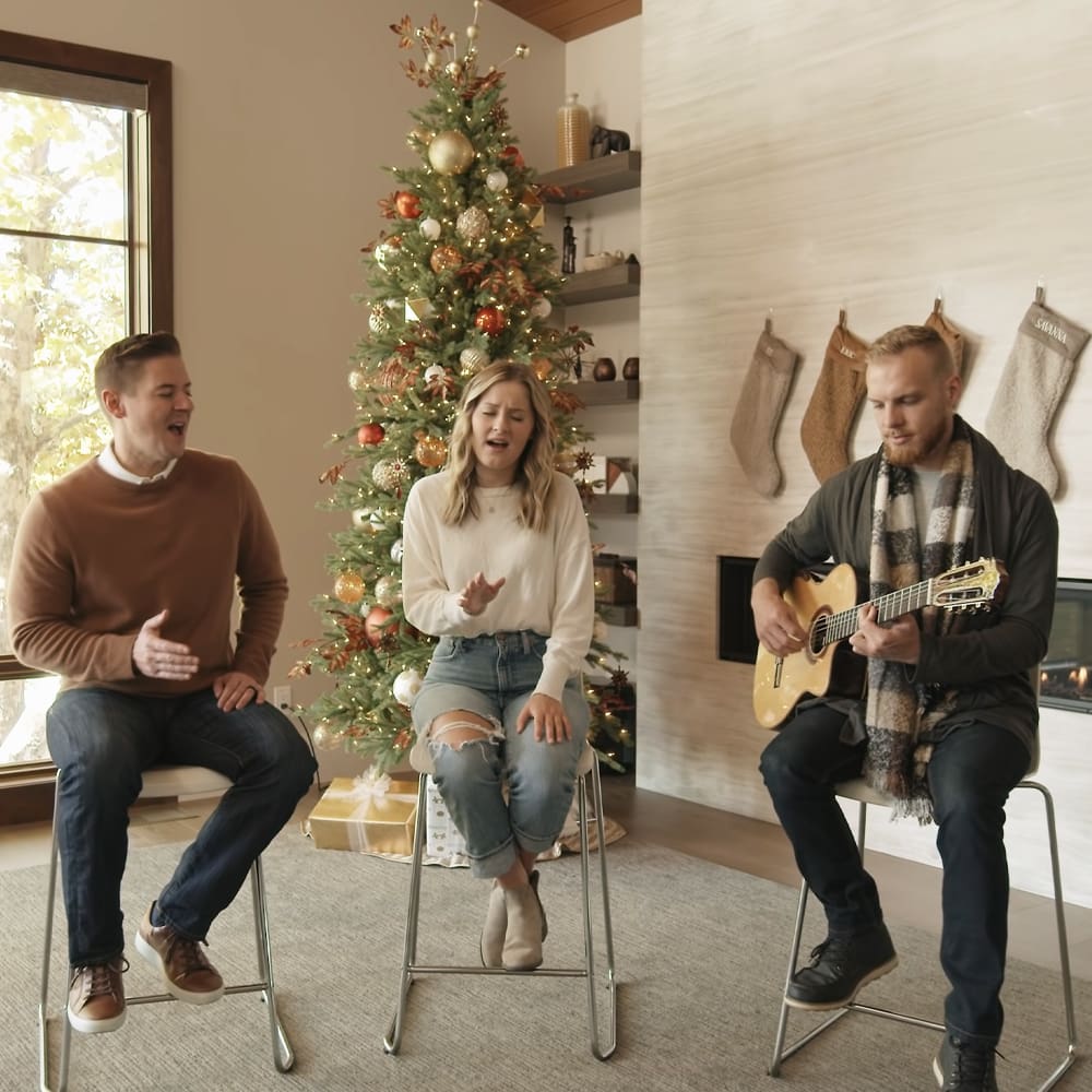 A family of three in a cozy living room decorated for Christmas with a fireplace, stockings, and a decorated tree.