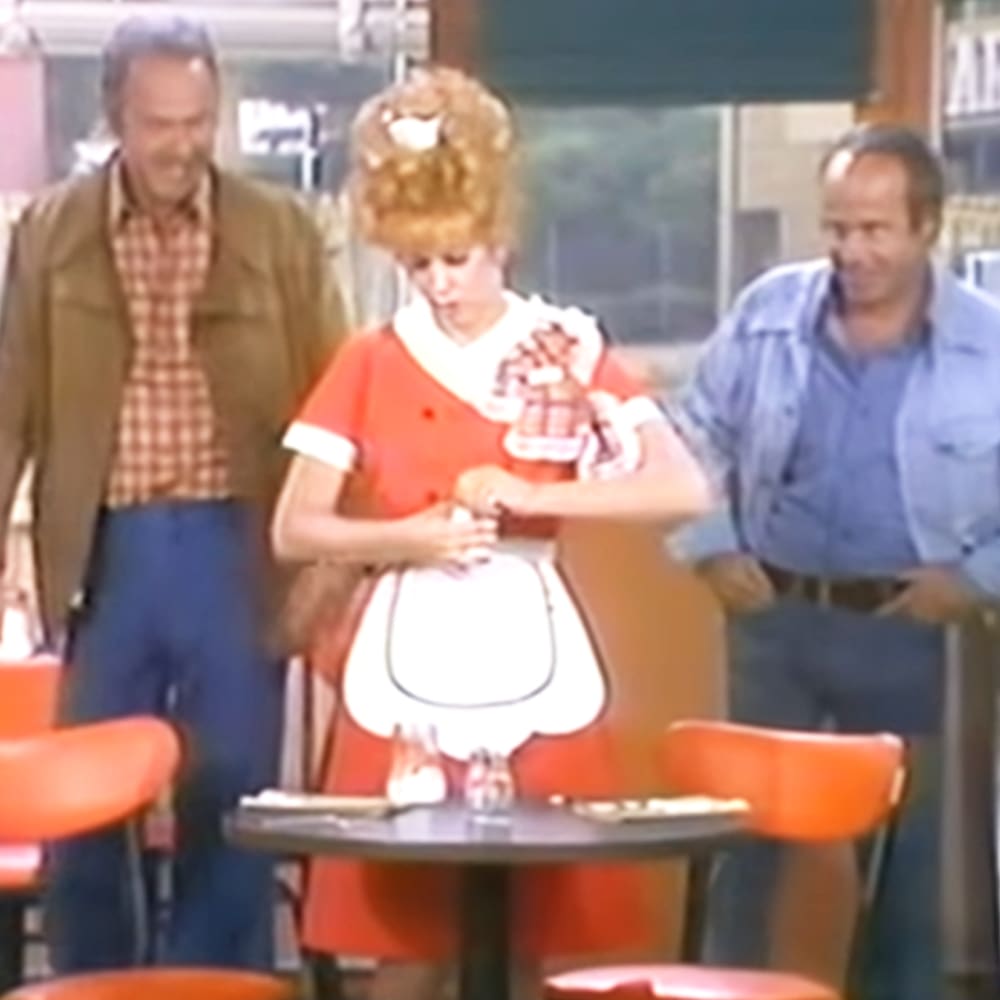 A young adult female in a diner, wearing a white apron with a circular cut-out design and an orange shirt. She is surrounded by other diners in casual attire, with visible furniture including black tables and orange chairs.