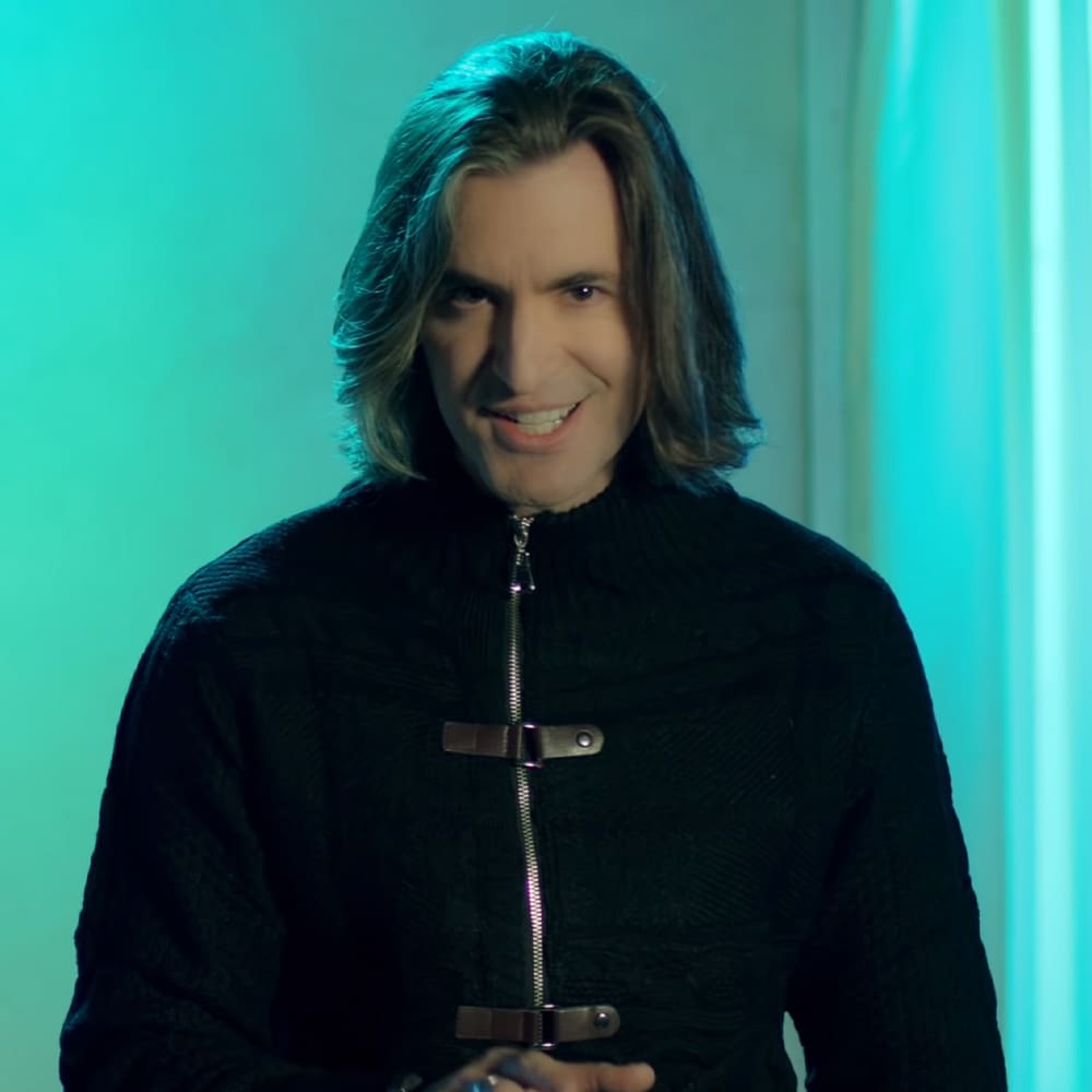 A middle-aged adult male wearing a black zip-up jacket with a textured pattern and metal zipper, set against a teal background under studio lighting.