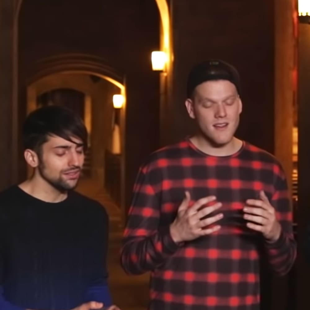 A young adult male wearing a dark blue long-sleeve shirt stands indoors with another person in a checkered sweater. The setting is an evening hallway with warm lighting and arched doorways.