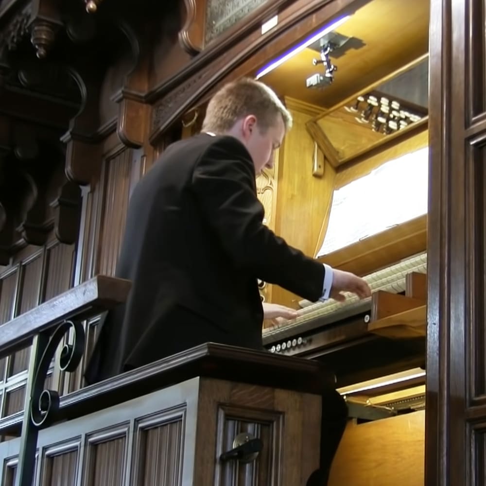 A young adult male playing an organ in a church or concert hall with ornate wooden paneling and decorative carvings, well-lit by interior lighting fixtures.