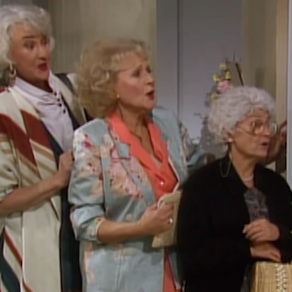 An older adult female in a white and red striped jacket with black trim, an older adult female in a light blue floral-patterned jacket over orange attire, and another older adult female in a textured black top or dress stand indoors against a neutral-colored wall.
