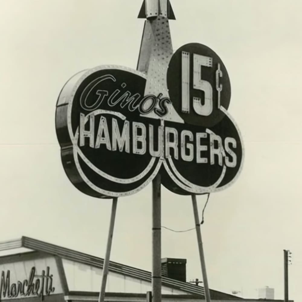 A large sign reading 'Gino's Hamburgers 15¢' in an urban setting with clear sky and daytime lighting.