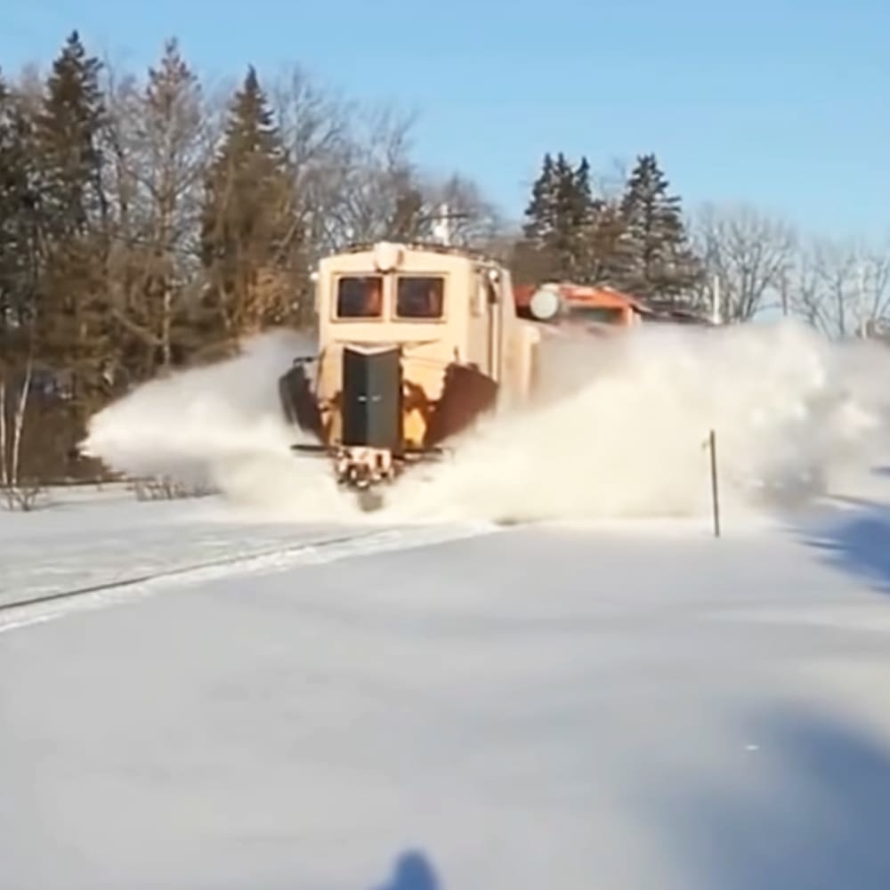 A snowplow clears a road in an outdoor winter scene with trees and bright sunlight casting shadows.