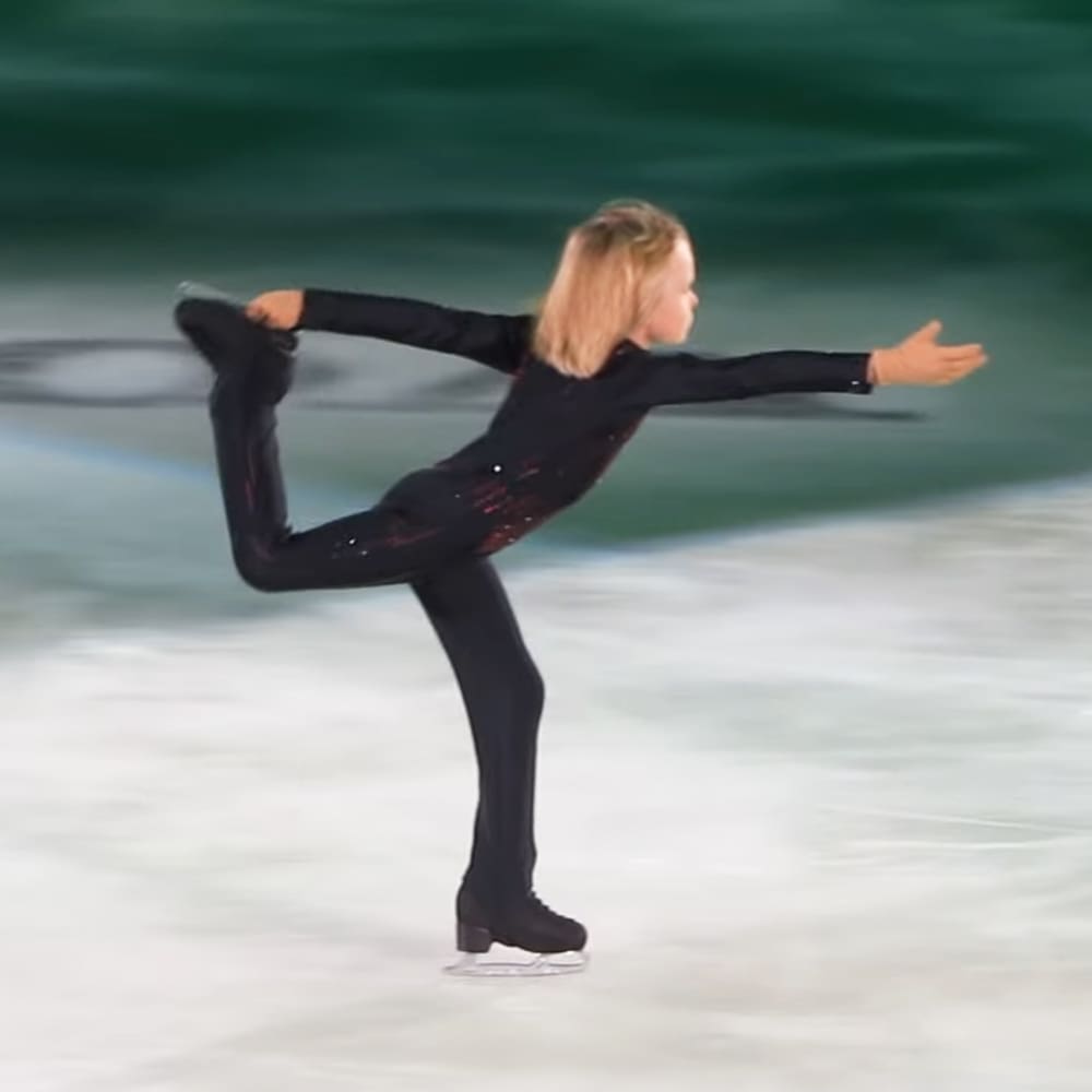 A young female figure skater in a black outfit with red accents, practicing at an indoor ice rink under artificial lighting.