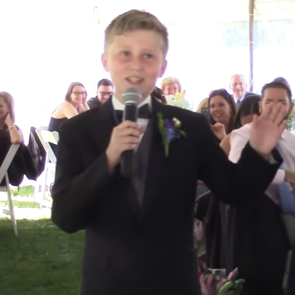 A young boy wearing a black tuxedo with a white shirt and purple boutonniere at an outdoor wedding ceremony under natural daylight.