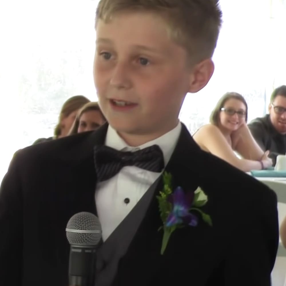 A young boy wearing a black tuxedo and purple boutonniere stands at a microphone in a formal indoor setting with adults seated around tables.