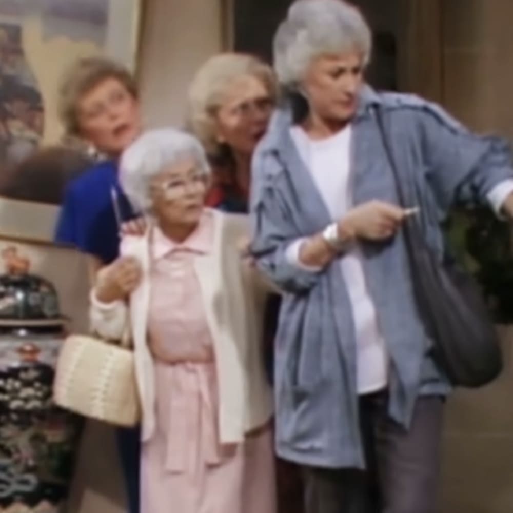 A group of older adults indoors, possibly in a home or community center; one woman in light pink dress with white cardigan and beige handbag, another pointing at something, others carrying bags or no items visible.
