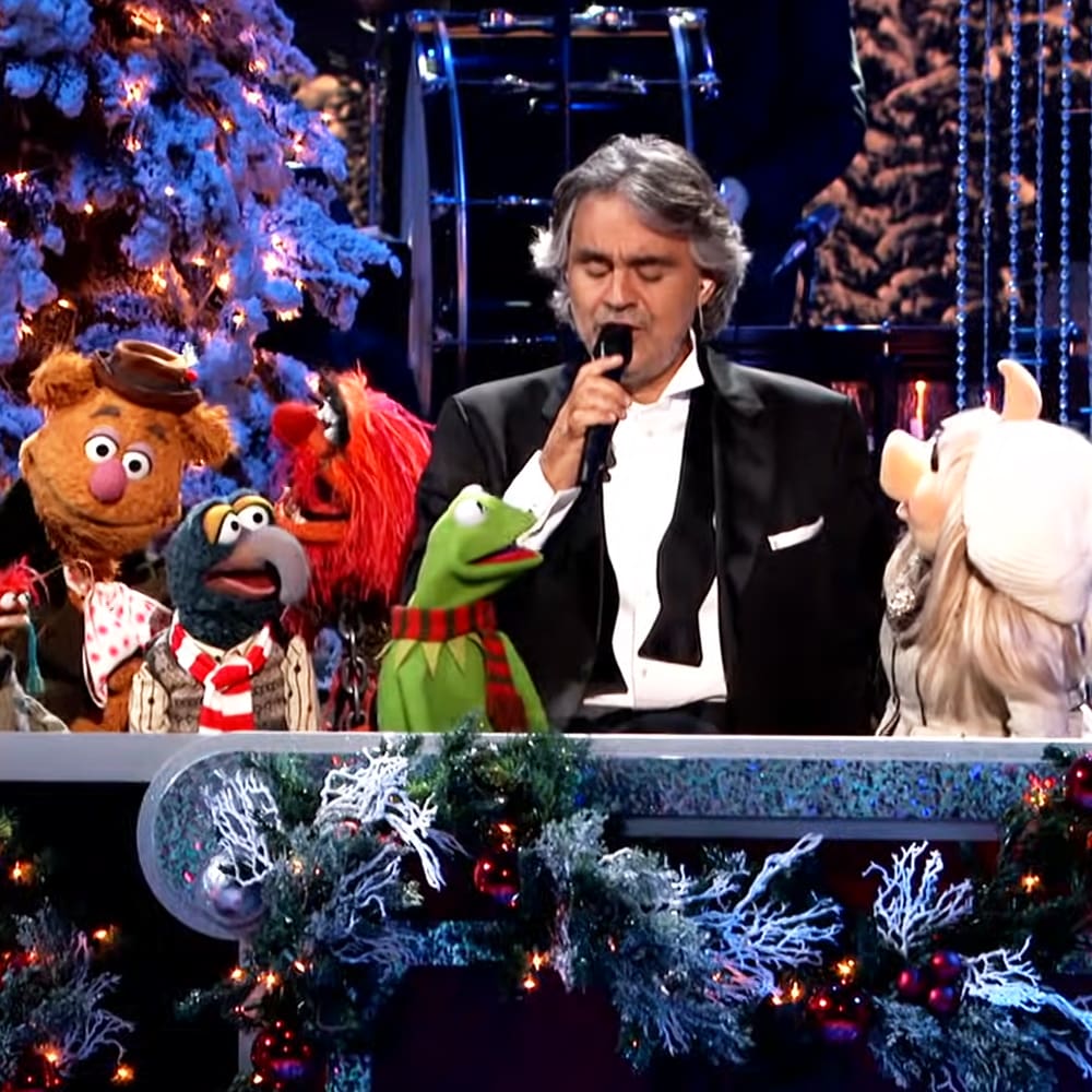 Andrea Bocelli in a black tuxedo jacket performing at a Christmas event alongside Fozzie Bear, Rowlf the Dog, Kermit the Frog, and Miss Piggy. Festive decorations and musical equipment are visible on stage.
