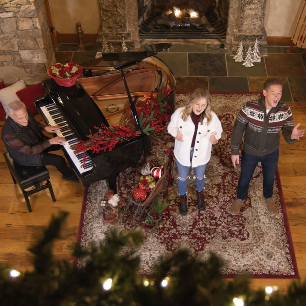 A family sings in a cozy living room decorated for Christmas, with a grand piano, fireplace, and ornate rug.