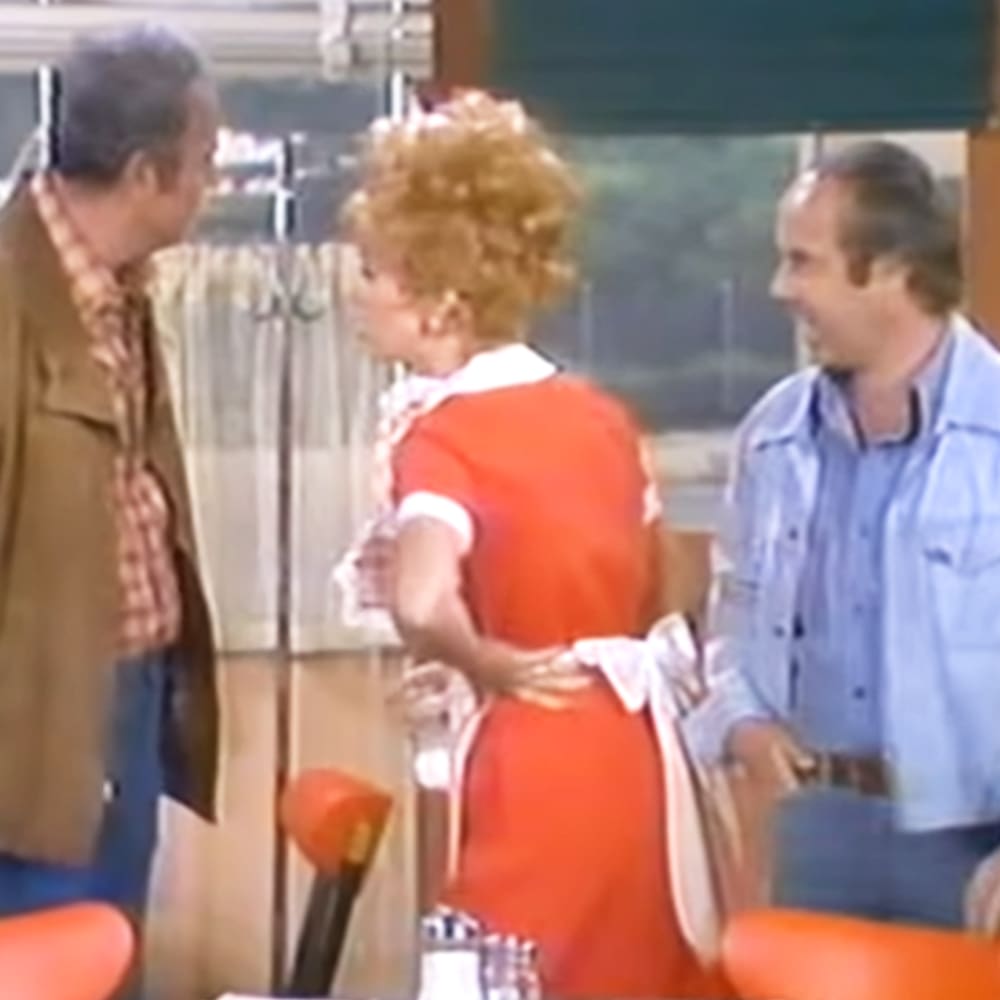 A woman in an orange dress with white trim stands between two men, one wearing a brown jacket over a plaid shirt and dark pants, the other in a light blue button-up shirt and dark pants. The setting appears to be indoors, possibly in a home dining or living room.