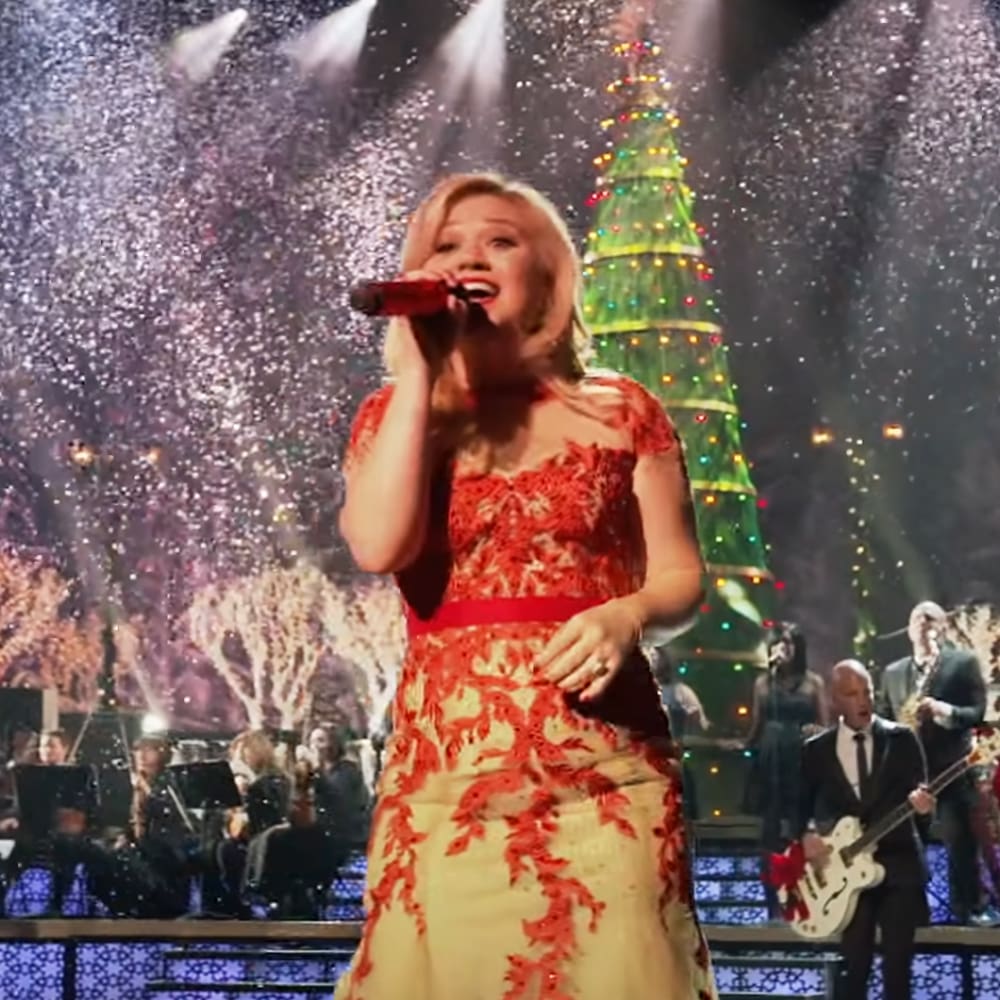 A female performer in a red and cream floral dress sings at a holiday-themed concert with an audience wearing dark suits, with festive decorations like a Christmas tree.