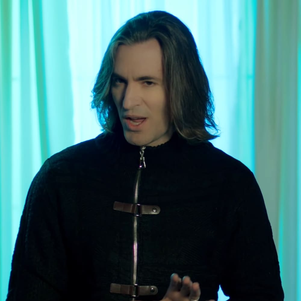 A young adult male wearing a black zip-up jacket with high collar and buckles, standing indoors under blue-green vertical light strips.