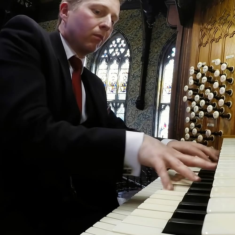 Adult male playing an organ inside a church with stained glass windows, formal attire suggesting the 20th century or early 21st century.