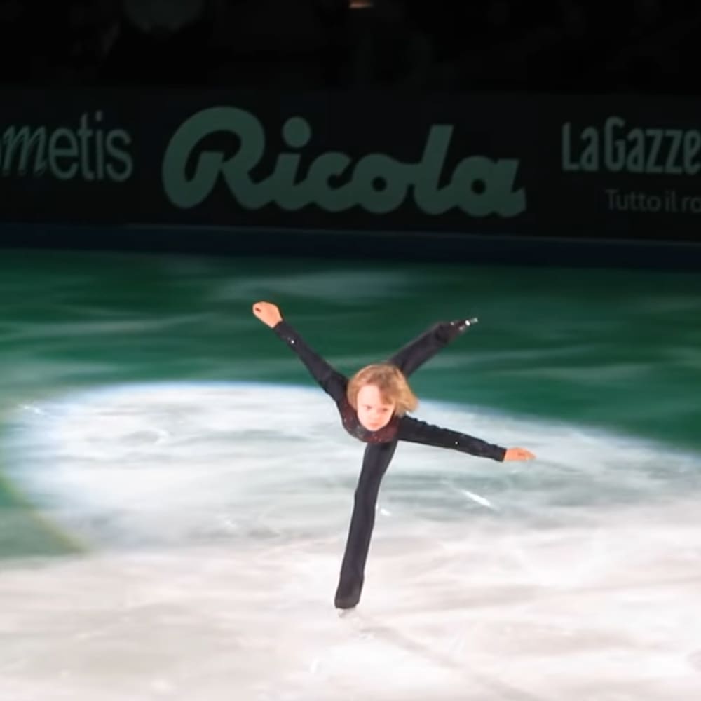 A young figure skater in a black outfit performs a spin on an indoor ice rink with advertisements and sponsor signs visible.
