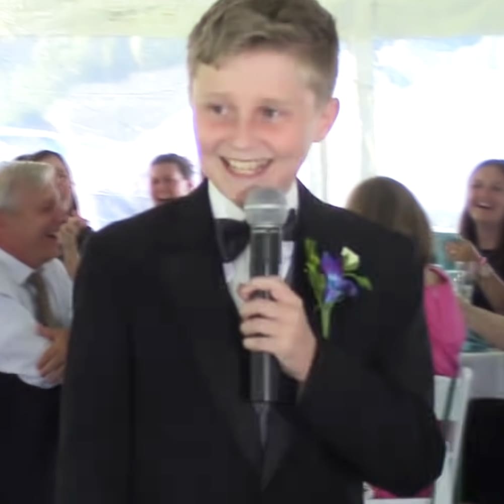 A young boy in a black tuxedo with a white shirt and bow tie, standing amidst adults at what appears to be an outdoor formal event under tent-like structures. The setting includes white plastic chairs and natural daylight.