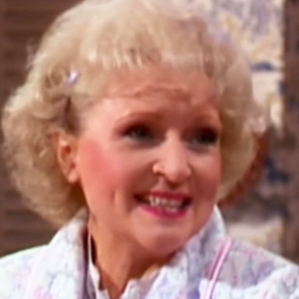 An older adult female wearing a white floral-patterned garment with pink trim, standing indoors with soft natural light and warm tones in the background.