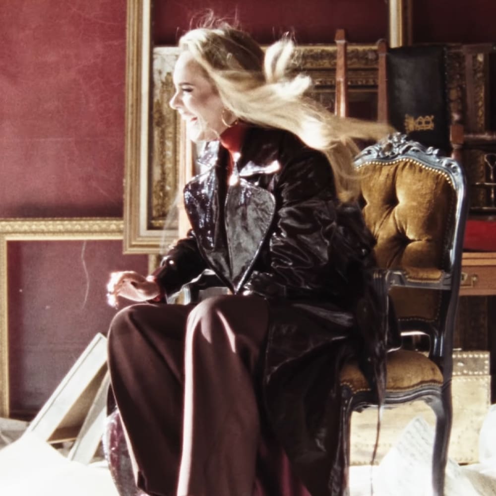 A young adult female wearing a shiny black leather jacket and dark brown pants in an indoor setting with ornate furniture, framed pictures on the wall, and red-painted walls.