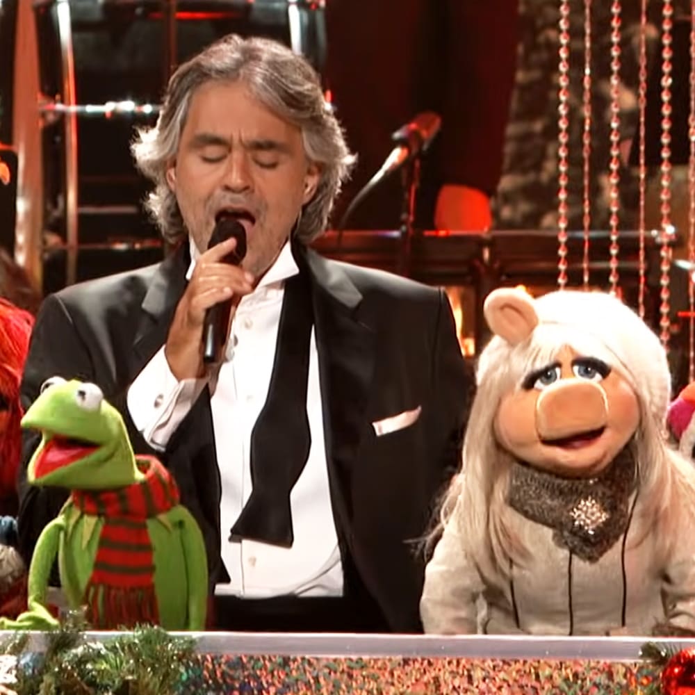 A middle-aged male singer in a black tuxedo performs on stage with Kermit the Frog and Miss Piggy puppets.