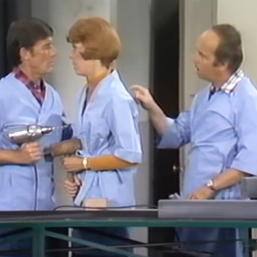 Indoor scene, possibly medical or laboratory setting; adults wearing light blue scrub tops with darker blue trim in relaxed fit, holding a metallic tool-like object; neutral-colored walls and door frame visible.