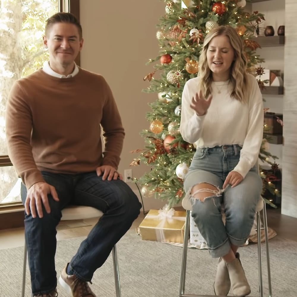 A young adult male and female in a festive Christmas setting indoors with decorated trees and holiday decor.