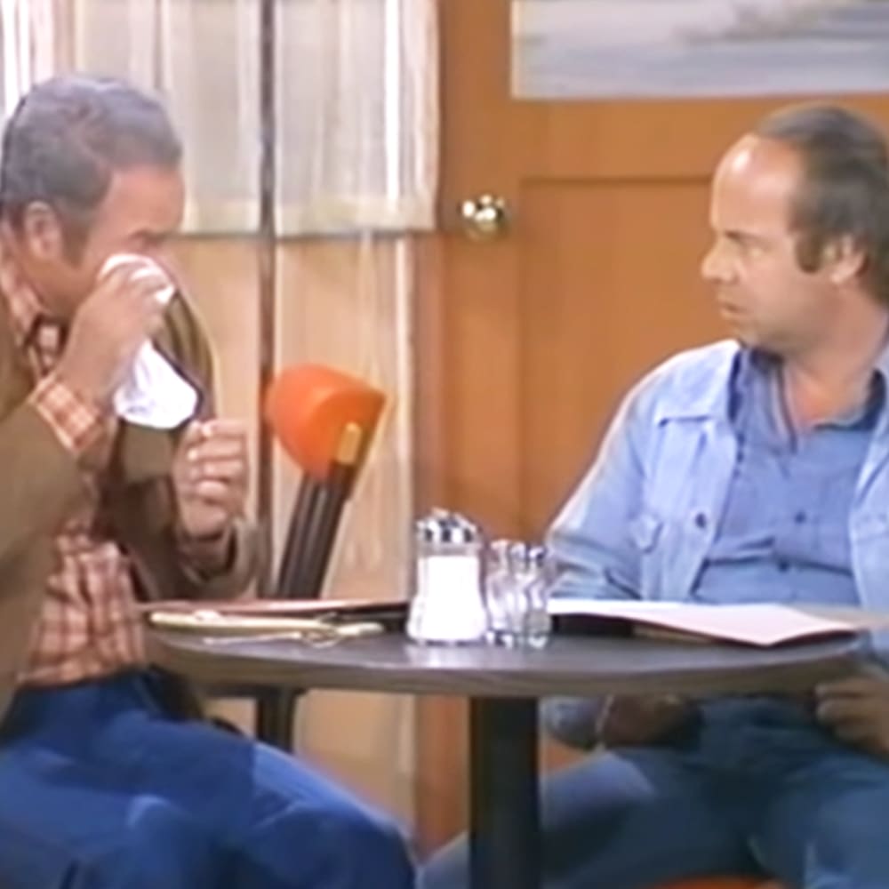 Middle-aged men in a casual dining scene with plaid shirts and light blue button-up shirts, seated around a dark round table with salt and pepper shakers and an open menu.