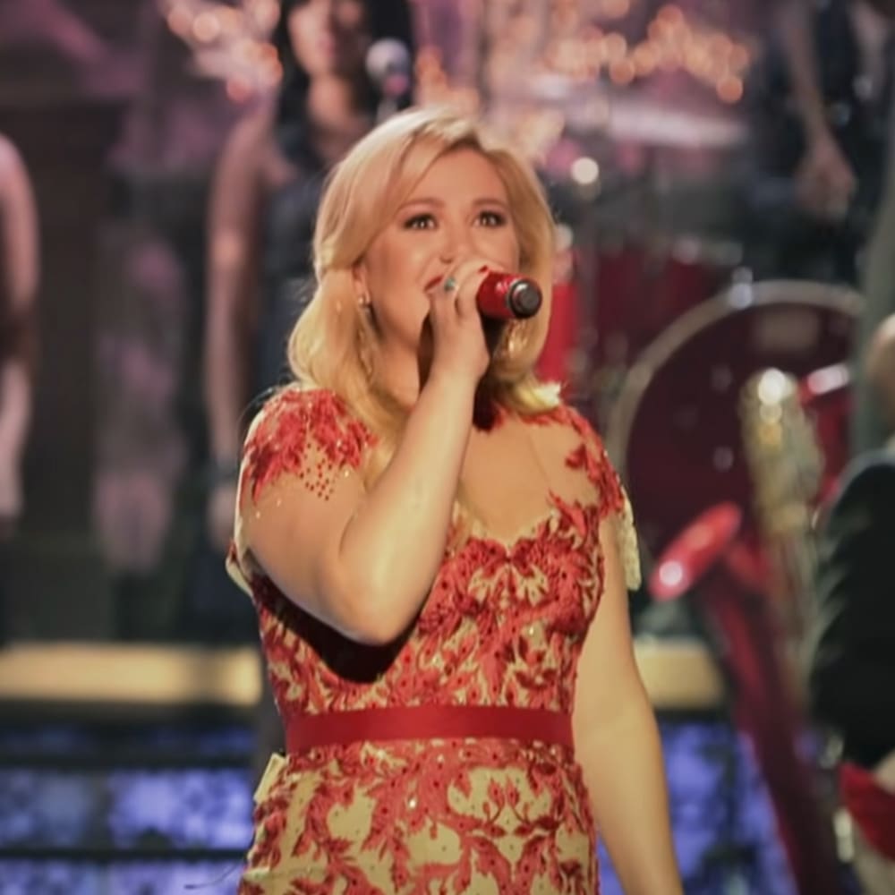A female performer singing into a microphone at an indoor concert, wearing a red and beige patterned dress with floral designs.